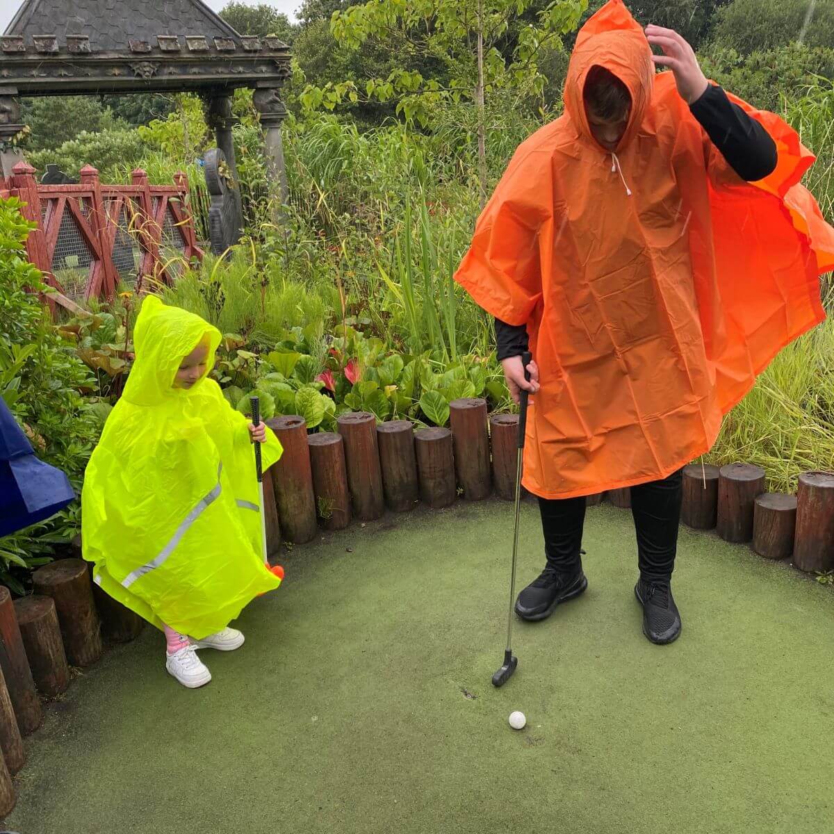 A man and child playing mini golf on holiday wearing rain macs.