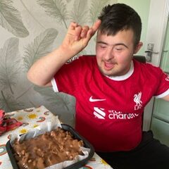Young man in a red t-shirt sat at a table with a tin of rocky road he has made.