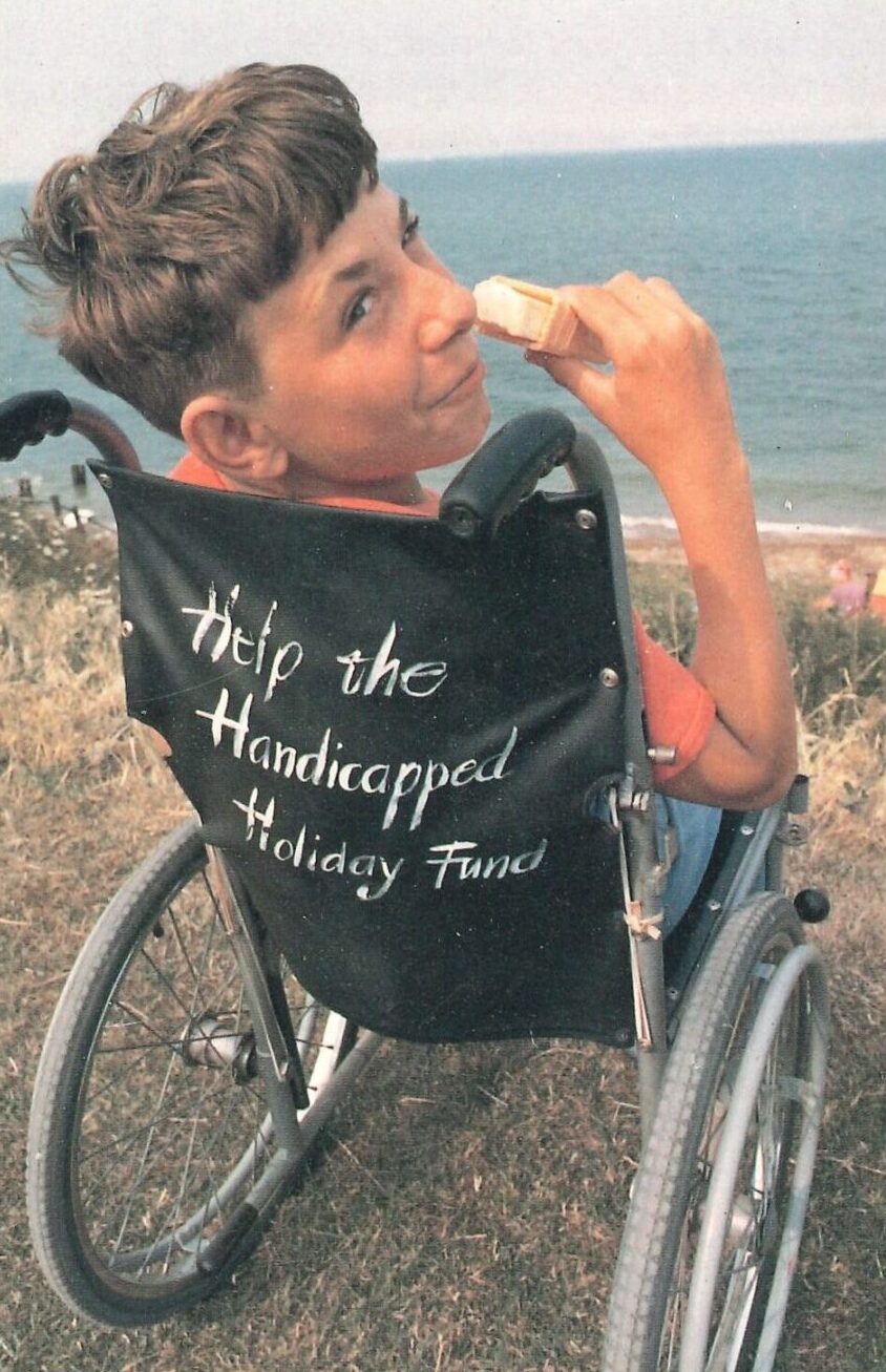 Boy in a wheelchair at the seaside looking over his shoulder eating an ice-cream.