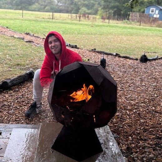 A man crouched next to a fire pit with countryside scenery behind him.