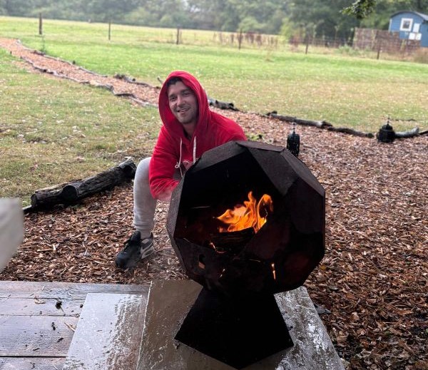 A man crouched next to a fire pit with countryside scenery behind him.