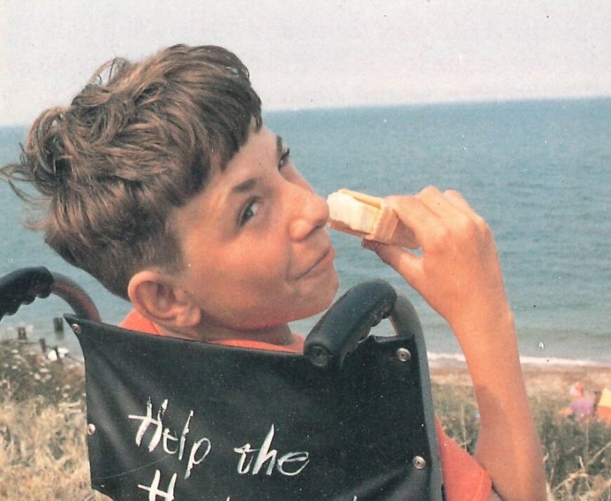 Young man with brown hair looking over his shoulder at the seaside while eating an ice-cream and sitting in a wheelchair.
