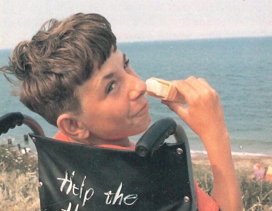 Young man with brown hair looking over his shoulder at the seaside while eating an ice-cream and sitting in a wheelchair.