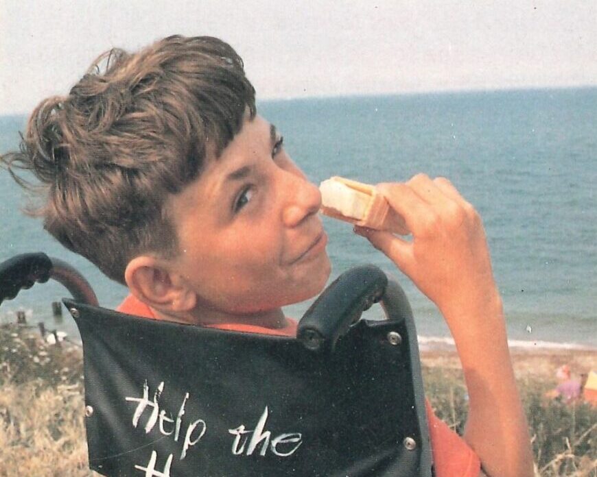 Young man with brown hair looking over his shoulder at the seaside while eating an ice-cream and sitting in a wheelchair.