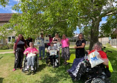 A group of men and women posing for the camera under a large tree with a farmhouse behind them. There is blue sky and bright sunlight. Some of the people are holding up 3H tote bags to show the camera.
