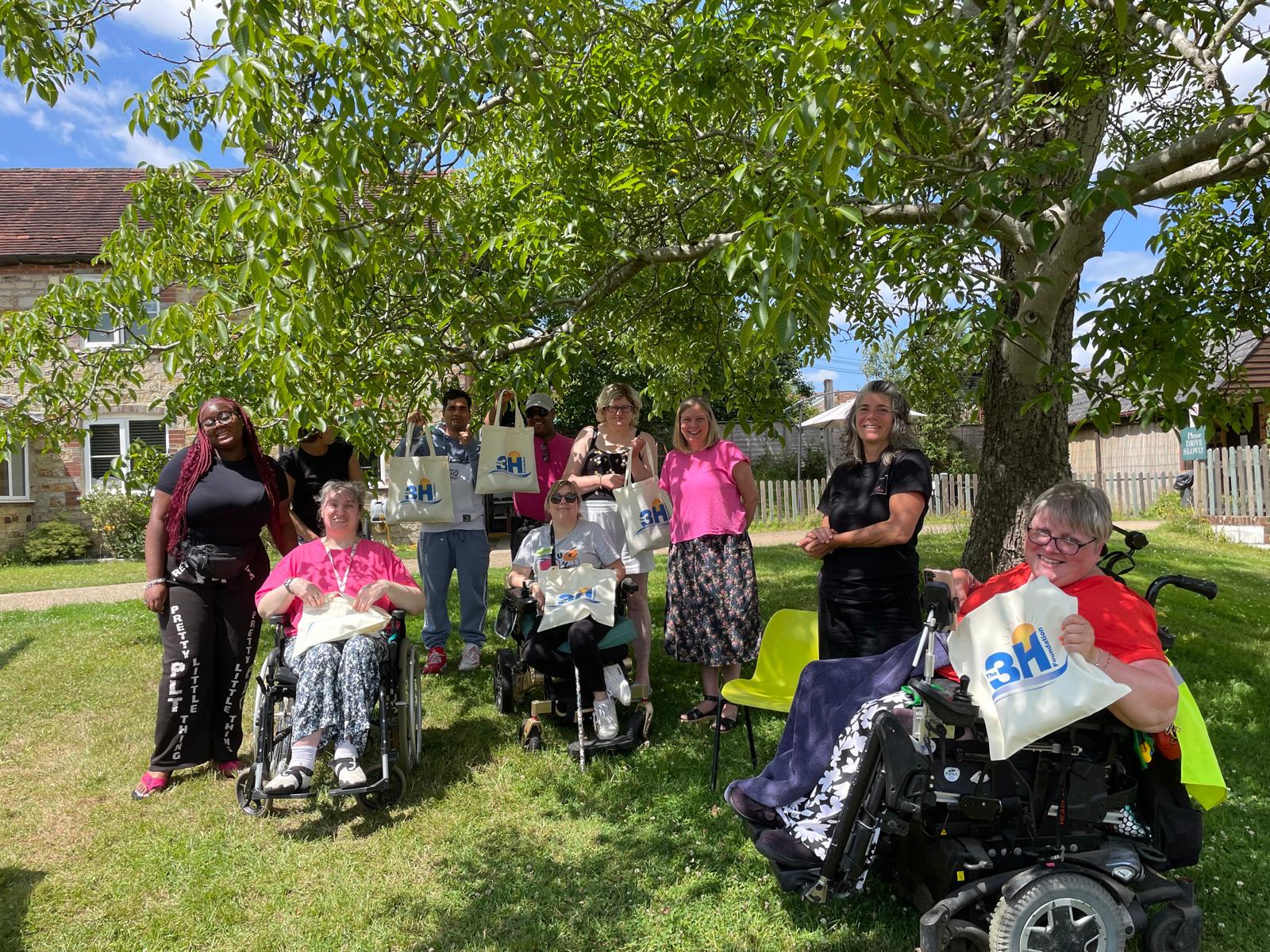 A group of men and women posing for the camera under a large tree with a farmhouse behind them. There is blue sky and bright sunlight. Some of the people are holding up 3H tote bags to show the camera.