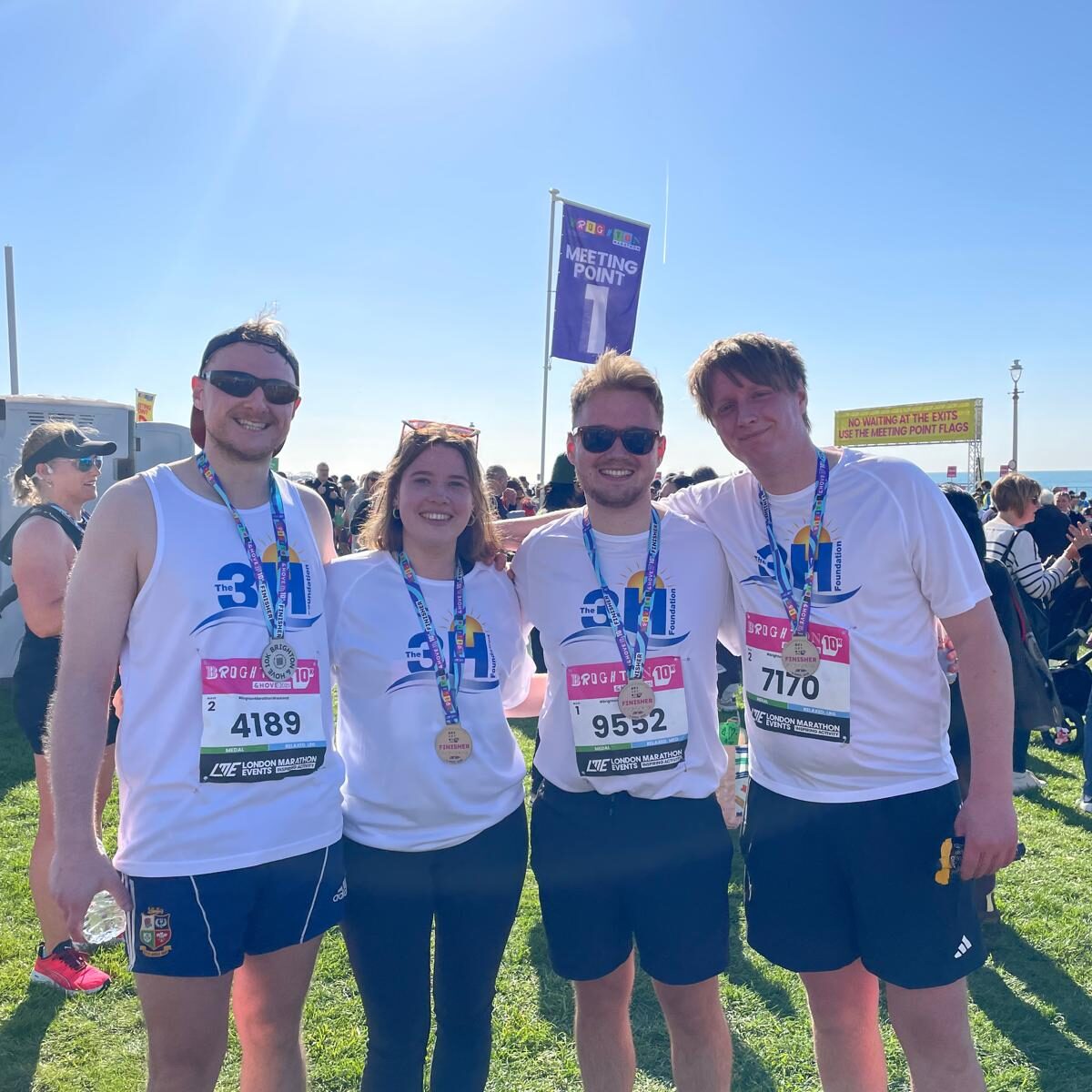 Brighton 10K 2025 Runners Four young adults facing the camera arm in arm. They are all wearing 3H running tops with their Brighton 10K numbers pinned to the front. The sun is out and the sky is blue behind them.