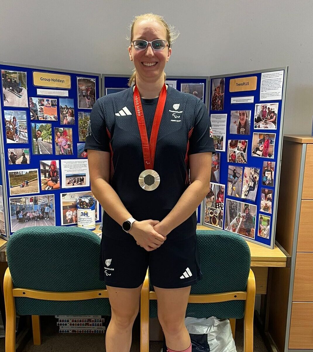 Callie-Ann Warrington standing in from of blue boards which have photos of beneficiaries enjoying their respite.