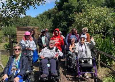 A group of men and women, some using wheelchairs and walkers, facing the camera. They are surrounded by trees and plants whilst on a path.