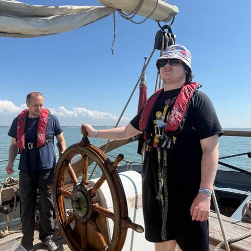 A young man wearing black and wearing a hat is steering a boat using a big wooden wheel. There is a man watching with a view of the sea and sky behind.