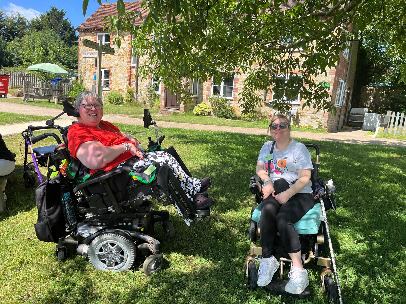 Two ladies sitting in wheelchairs in front of an old stone building smiling at the camera.  The sky is blue, the sun is out and they are on grass under a tree.