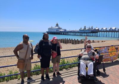 A group of men and women facing the camera while standing on a seaside promenade with blue skies, a shingle beach and a pier behind them.