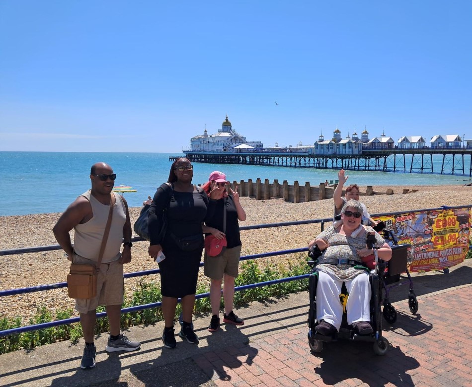A group of men and women facing the camera while standing on a seaside promenade with blue skies, a shingle beach and a pier behind them.