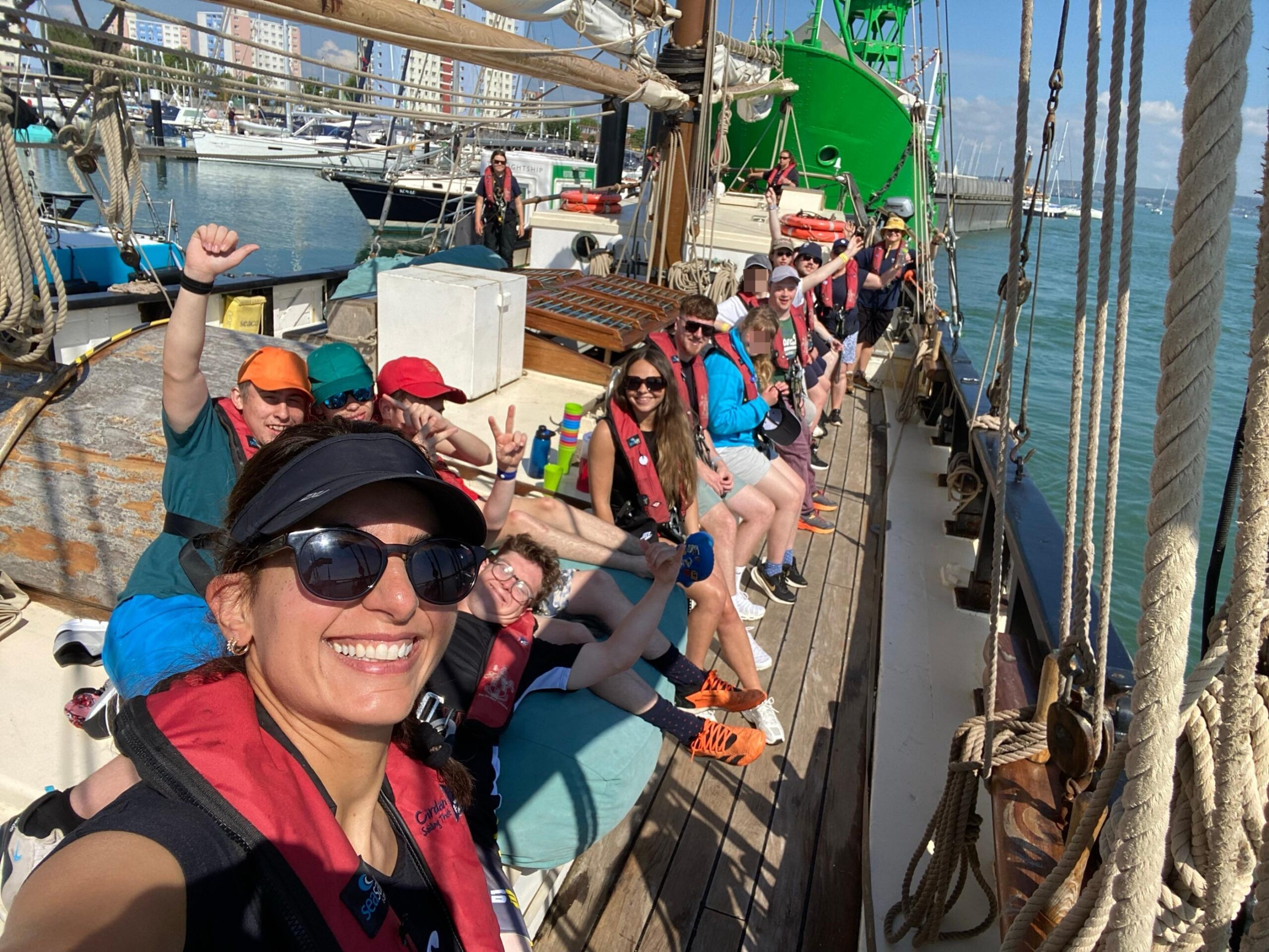 A group of people sitting on the deck of an old sailing boat looking at the camera.  while out at sea.