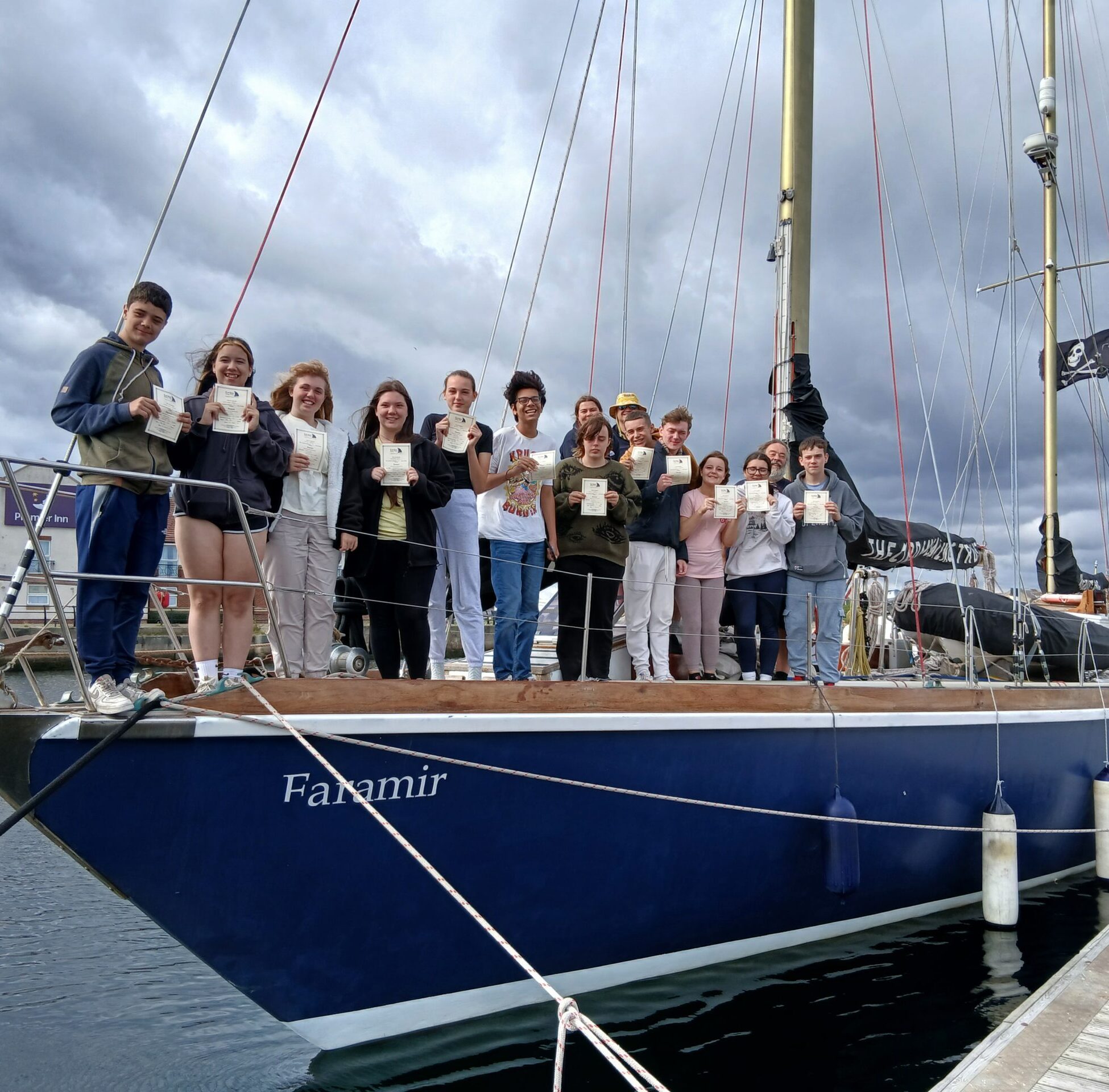 A group of young people standing on the deck of an old sailing boat proudly holding up their sailing certificates.