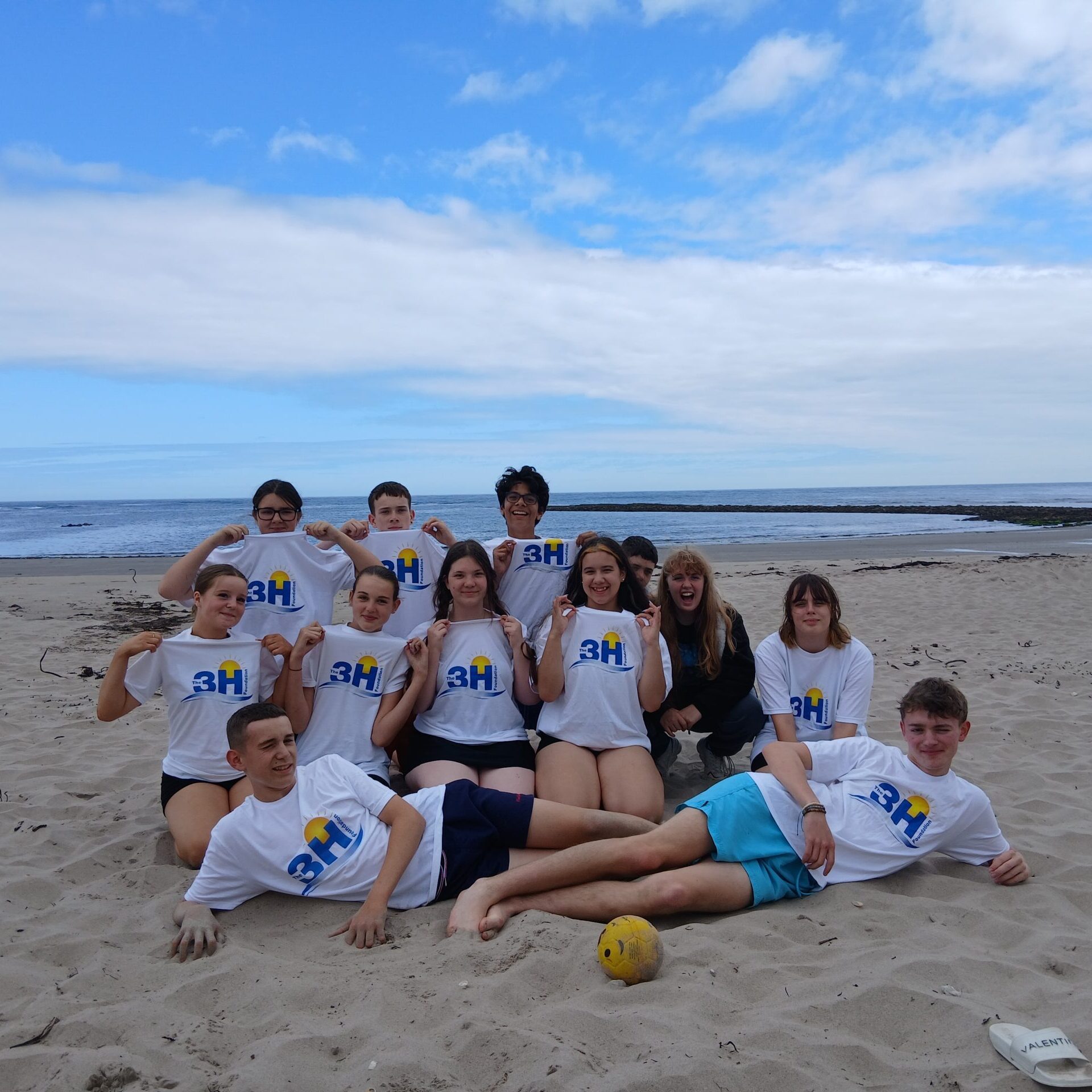 A group of teenage boys and girls sit on a beach in a group facing the camera with The 3H Foundation white t-shirts with the logo showing. The blue sky and sea is behind them.