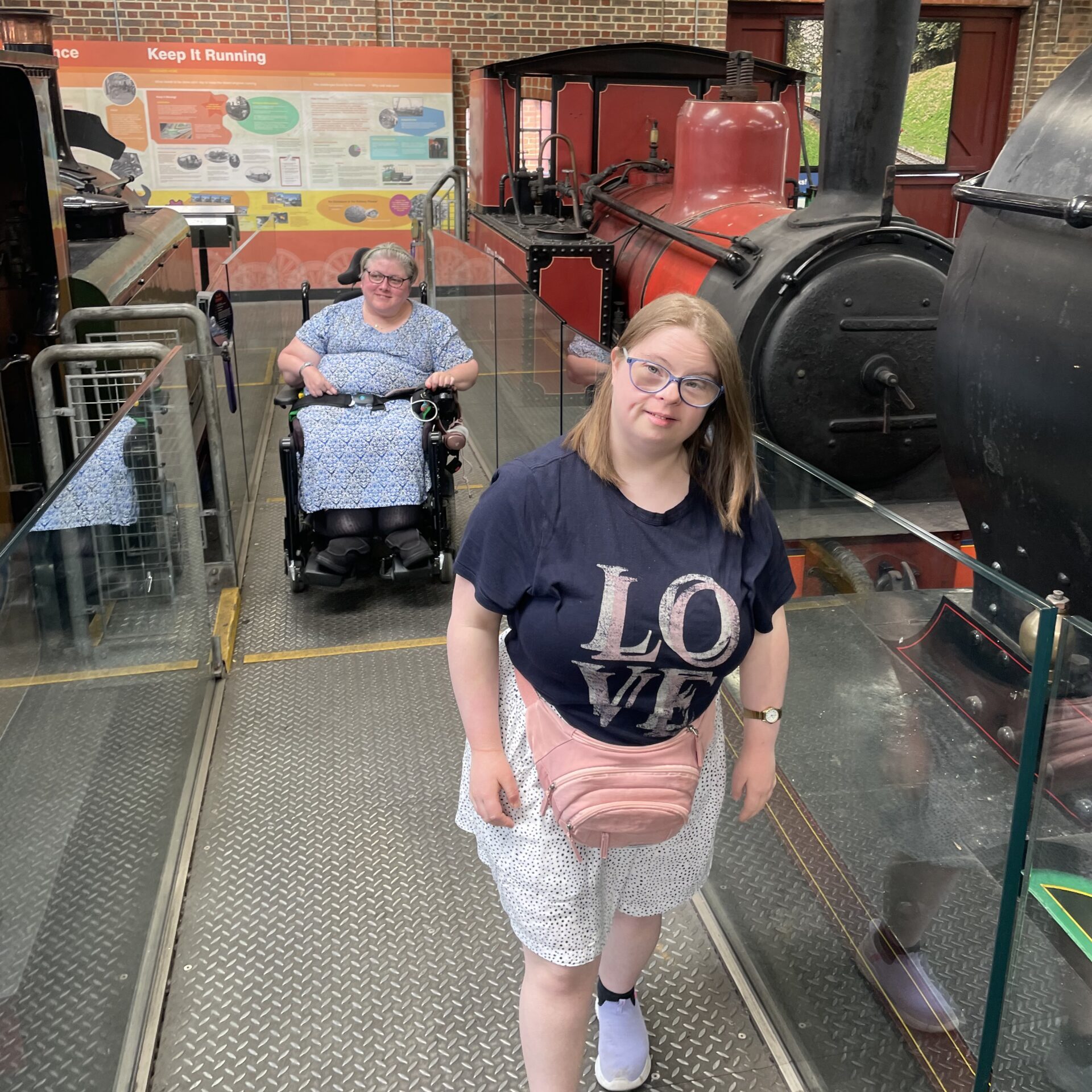 Railway Museum Two holiday guests both female inside a train museum looking at the camera. One young lady wearing a blue 'love' t-shirt and glasses is standing in front of another lady who is wearing a pale blue dress and is a wheelchair user.