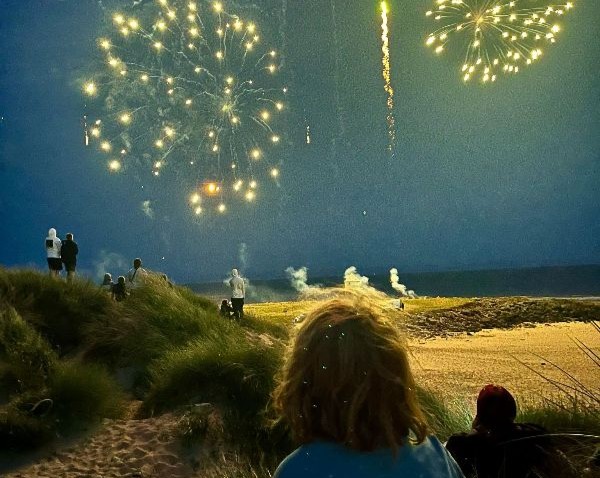 Two people sitting on a beach looking away from the camera towards fireworks in the sky over the beach.