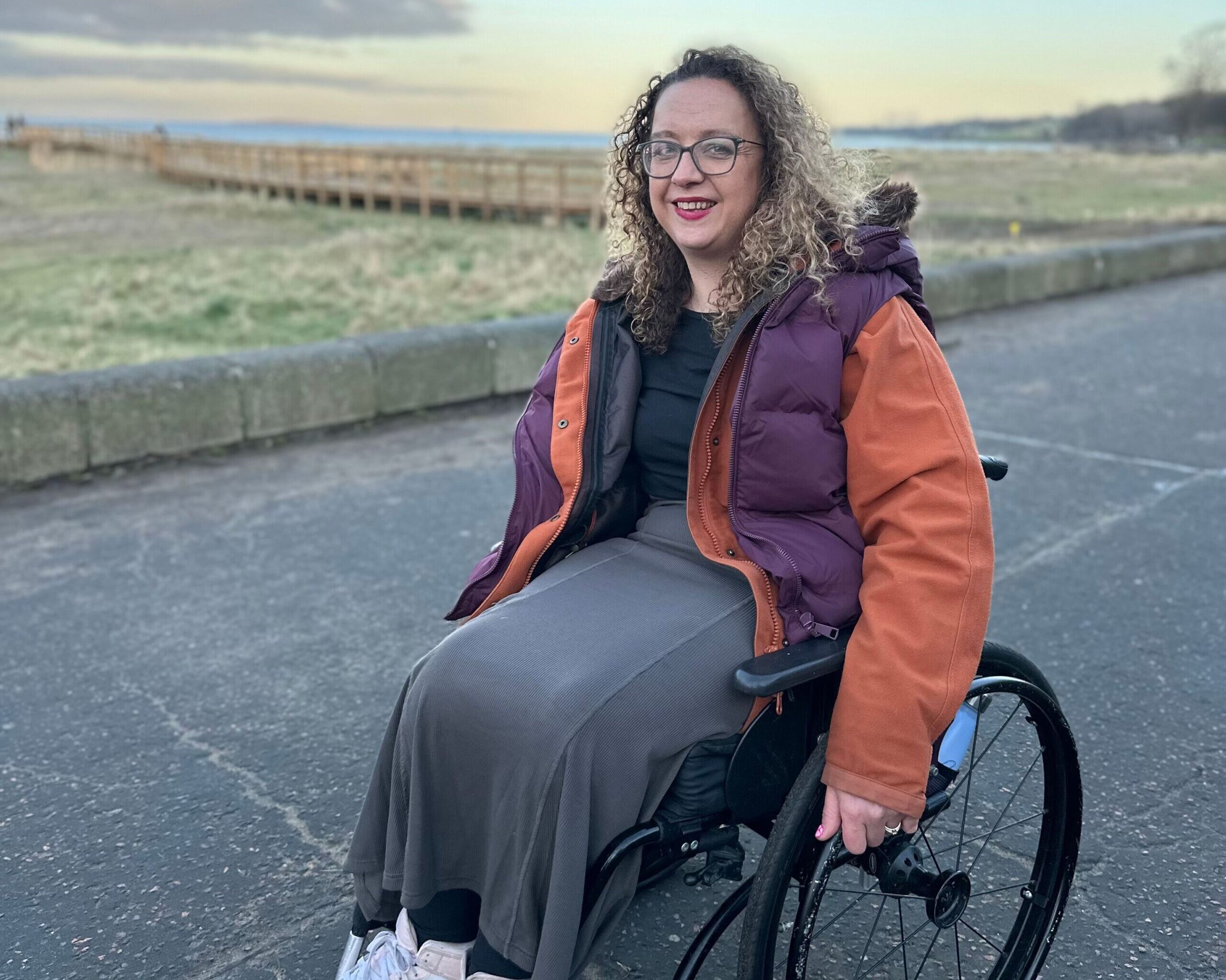 Carrie-Ann Lightley smiling at the camera with a view of sea, sky, grass and clouds behind her.