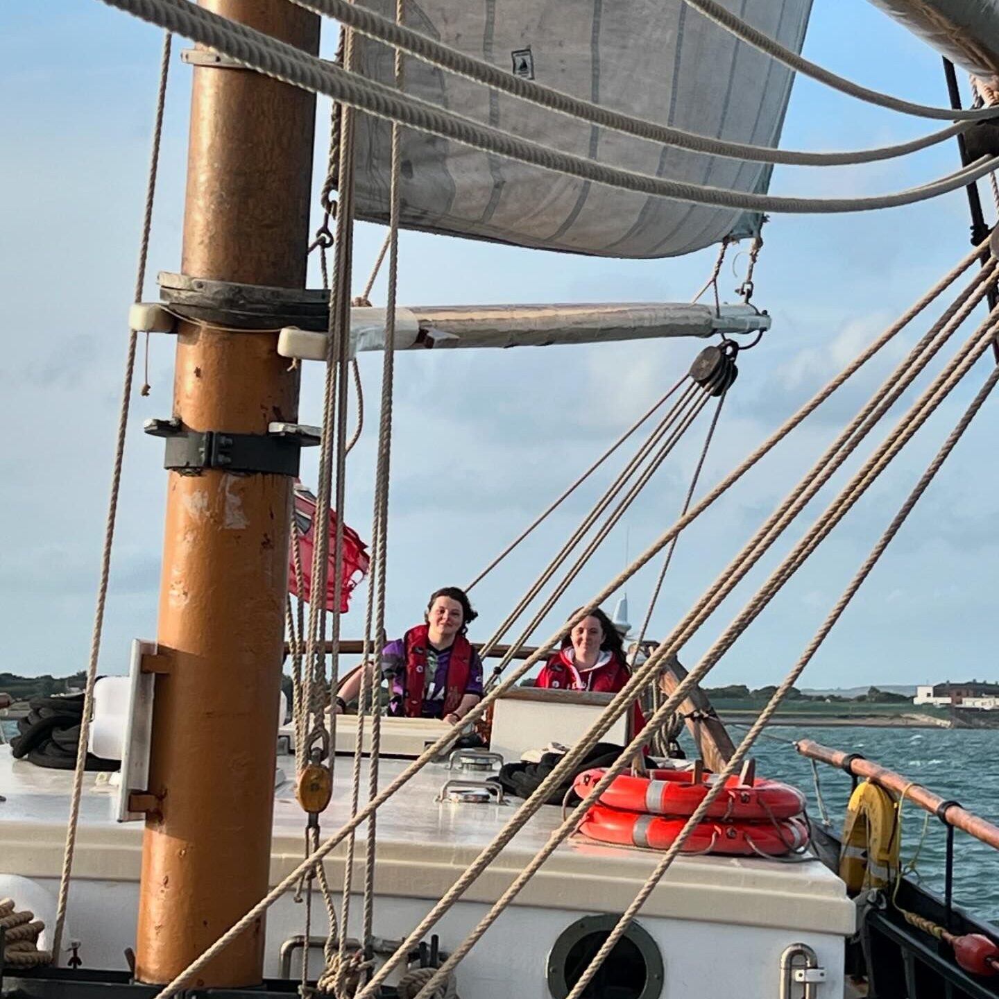 Two teenage girls are sailing a large sailing boat.  The photo was taken of the girls looking through the ropes and sail towards the steering wheel.