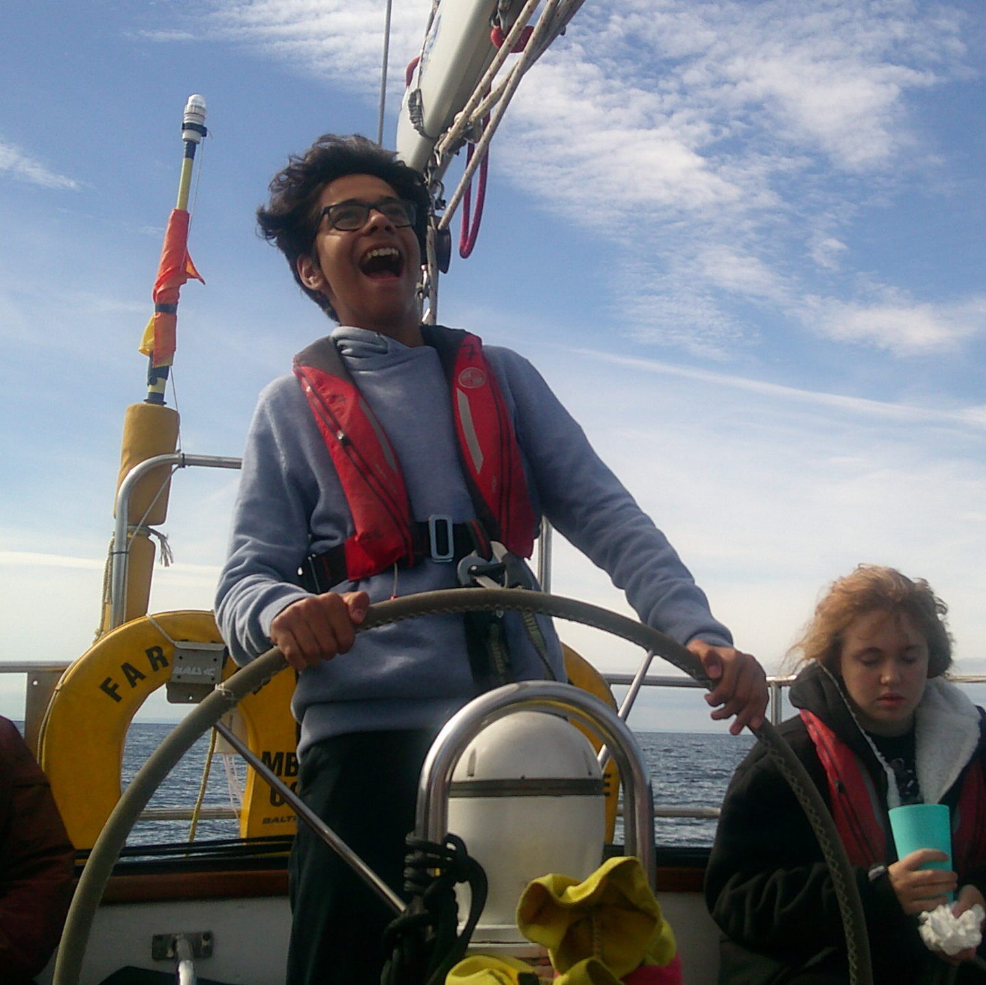 A young man stands at the helm of the Queen Galadriel sailing boat with the wind blowing his hair back and a big smile on his face.