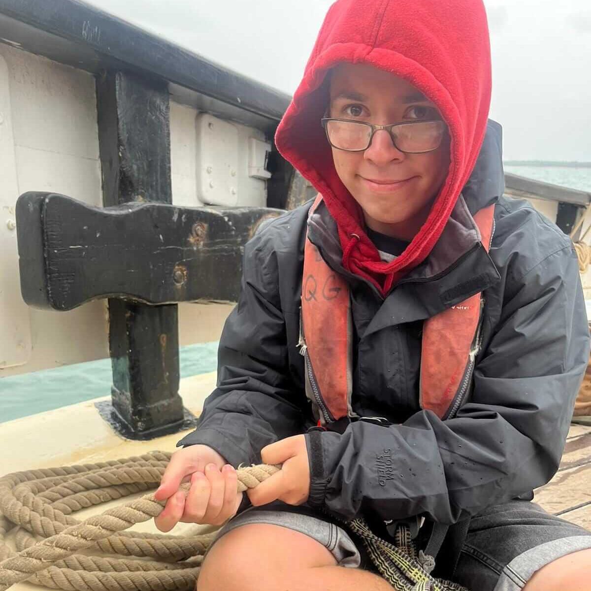 A boy wearing a red hooded top with the hood up under a black jacket sitting crossed legged on the deck of an old sailing boat. He is looking at the camera while holding some rope.