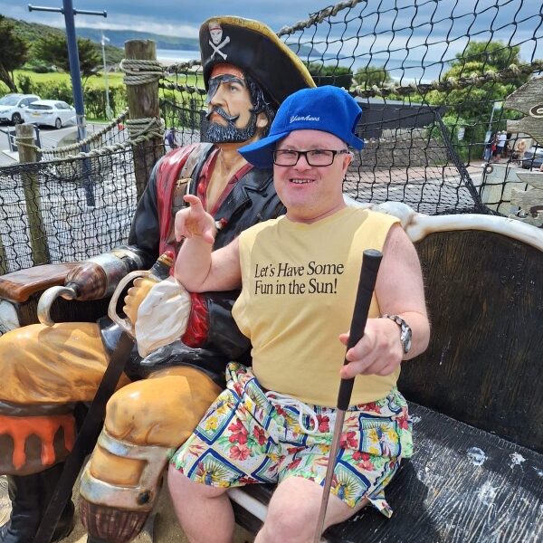 A young man is sitting beside a wooden life sized pirate. The man is wearing a blue baseball cap and yellow t-shirt. He is making the shape of a hook with his finger to copy the pirate hook.