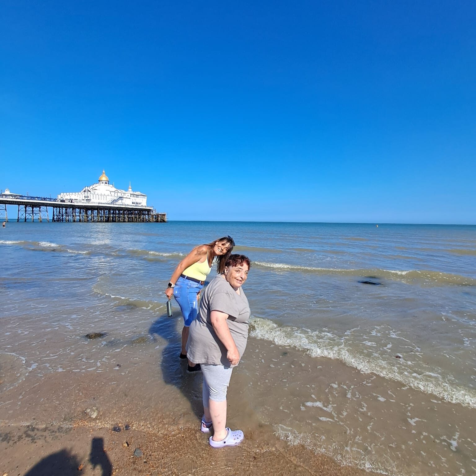 Two ladies paddling in the sea with the pier behind them.