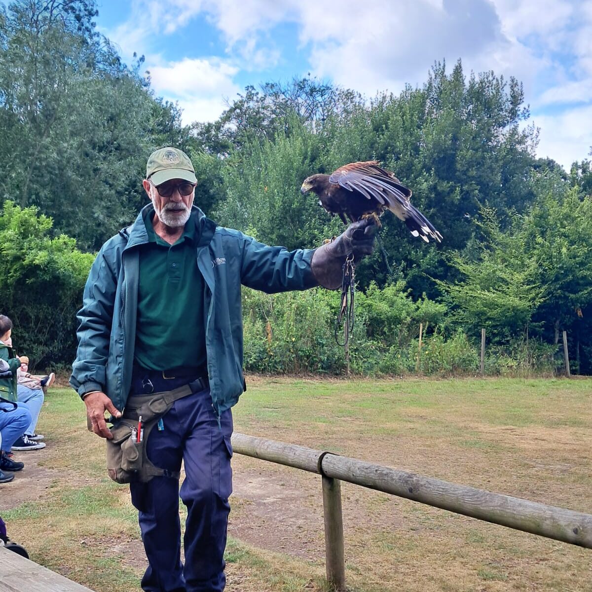 A man holding a large bird of prey on his hand while facing the camera. He is dressed in blue jeans, blue jacket and green jumper with a green baseball cap on.