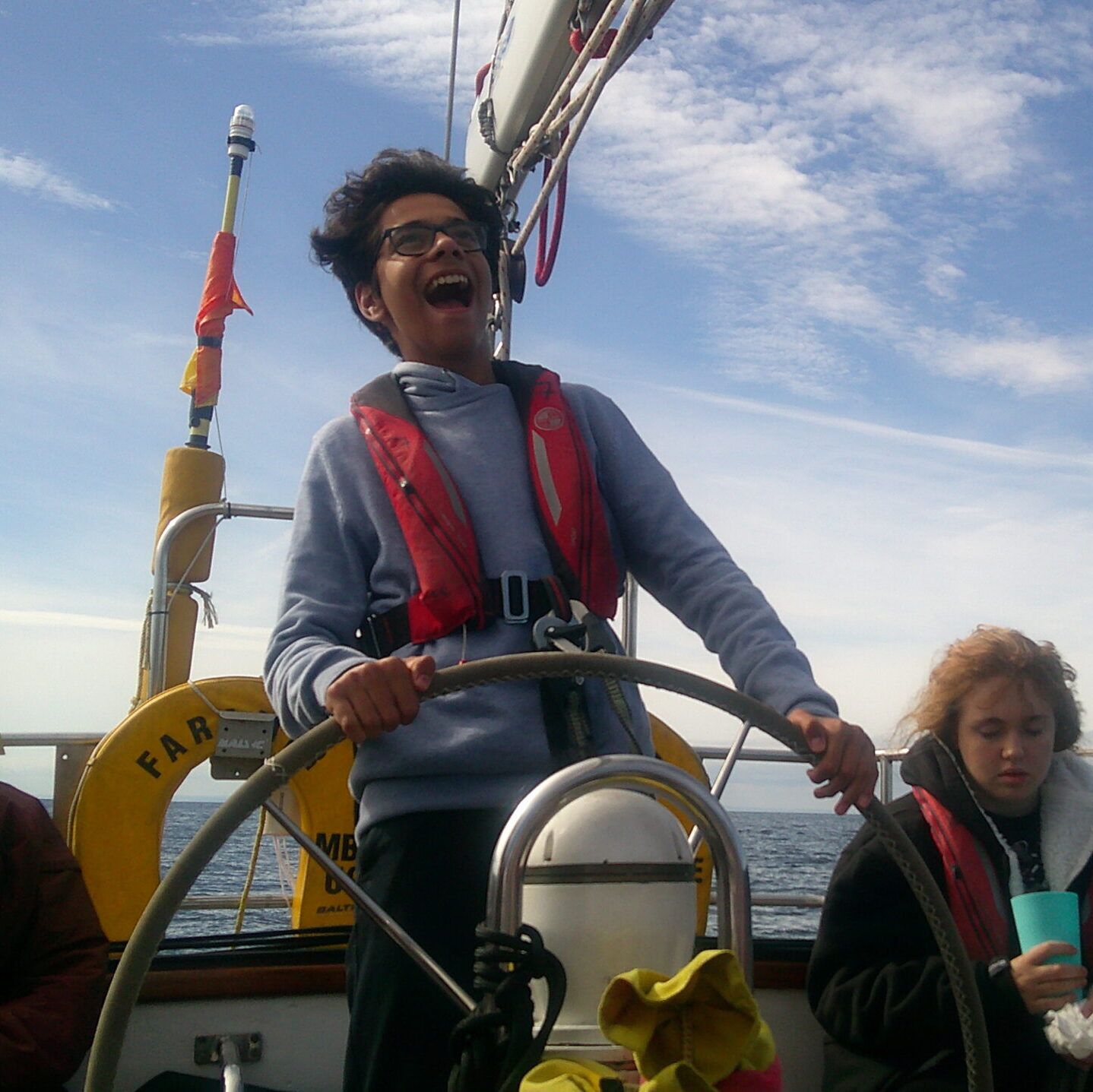 A teenaged boy is standing holding the wheel of the large sailing boat with the wind in his hair and a big smile on his face.