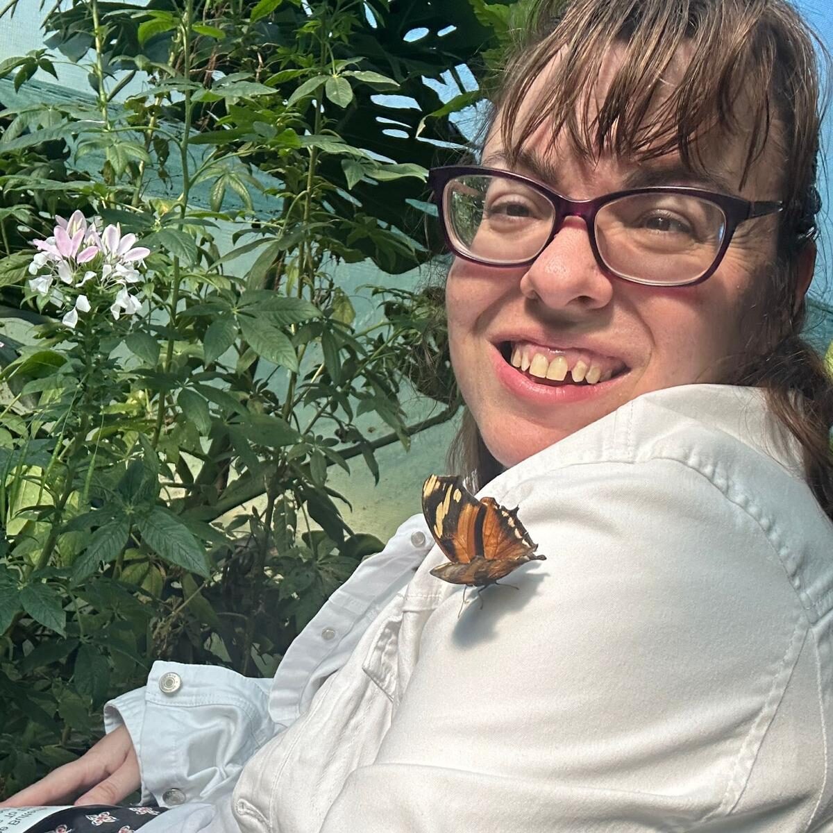 A young lady wearing a while top and glasses is smiling at the camera while looking over her shoulder. On her shoulder is a butterfly.