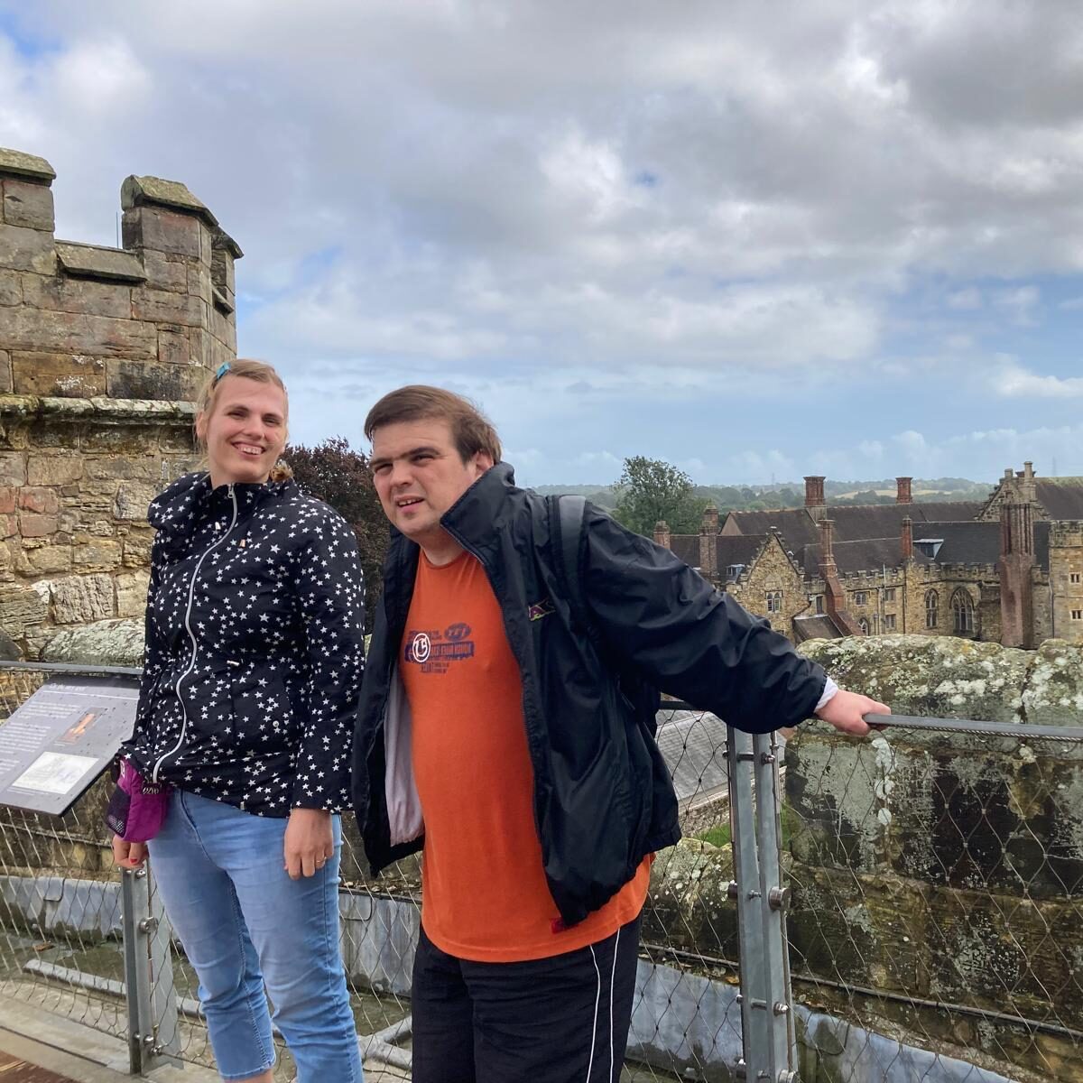 A man and a woman are standing at the top of an abbey looking at the camera with the view of old buildings behind them.
