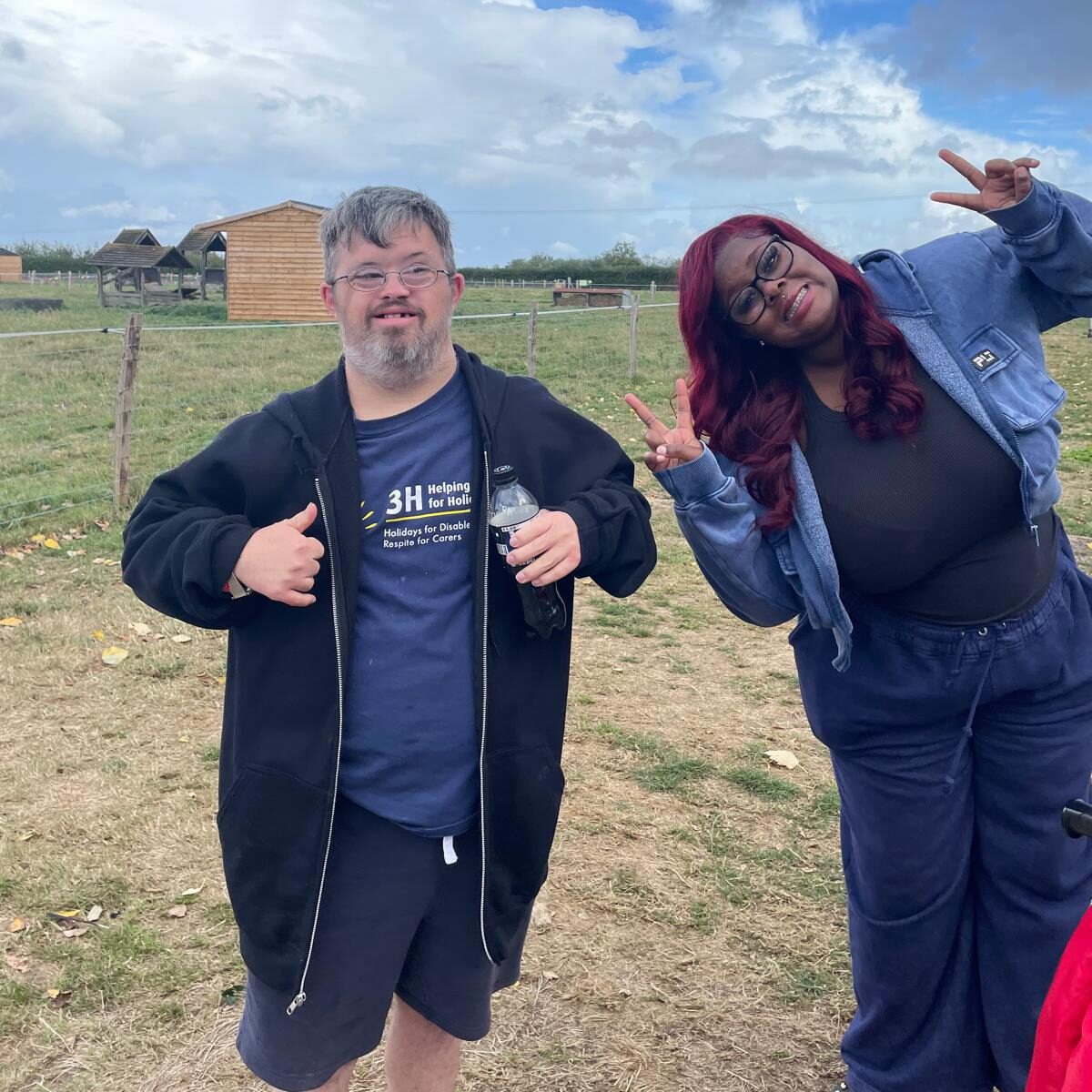 A man and woman are looking at the camera smiling. The man has his thumbs up and the woman has made peace signs with hers. They are standing in a field.