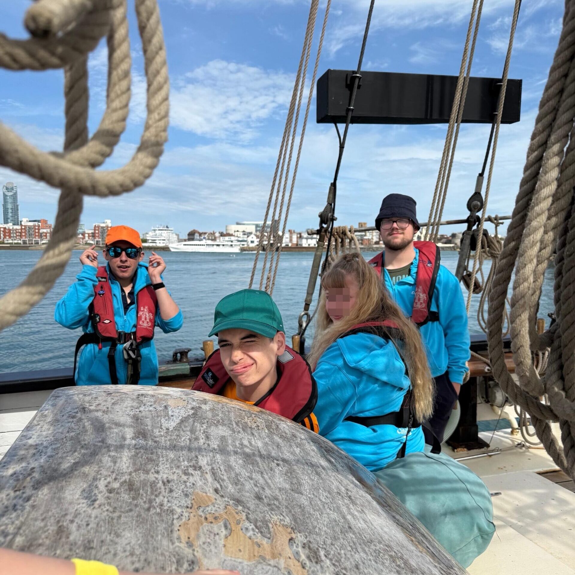 A group of young people on a large sailing boat looking at the camera with the port behind them.